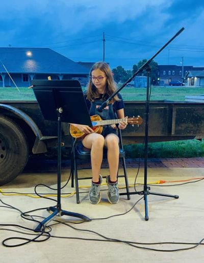 young girl seated on a stool behind a music stand with microphone performing on a ukulele