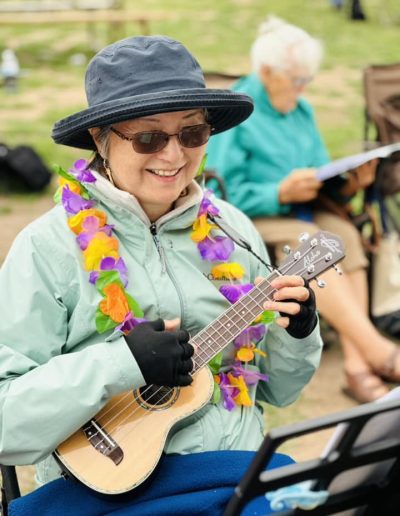 Older woman wearing fingerless gloves, jacket, hat, and a lei performing on a ukulele