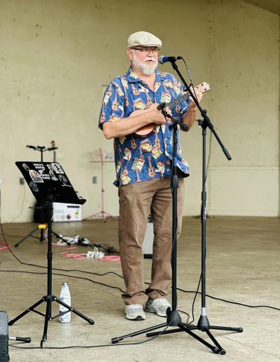 Man in a Hawaiian shirt standing behind a microphone on a stage performing on a ukulele