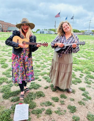 Two young women in skirts standing in a field in front of flags blowing in the wind performing on ukuleles
