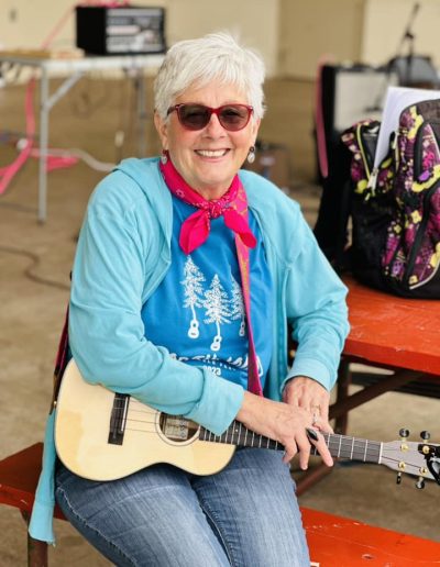 Older woman seated at a picnic table poses with a ukulele