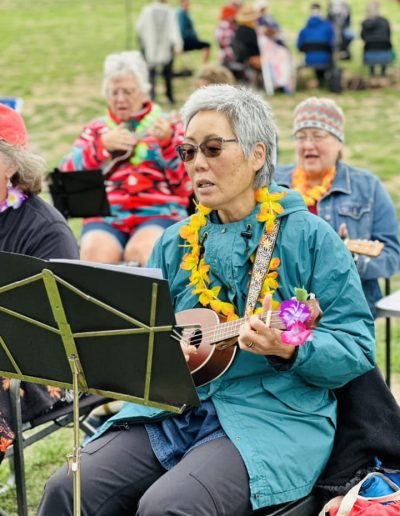 Older woman wearing a rain jacket sitting in field with other musicians performs on a ukulele