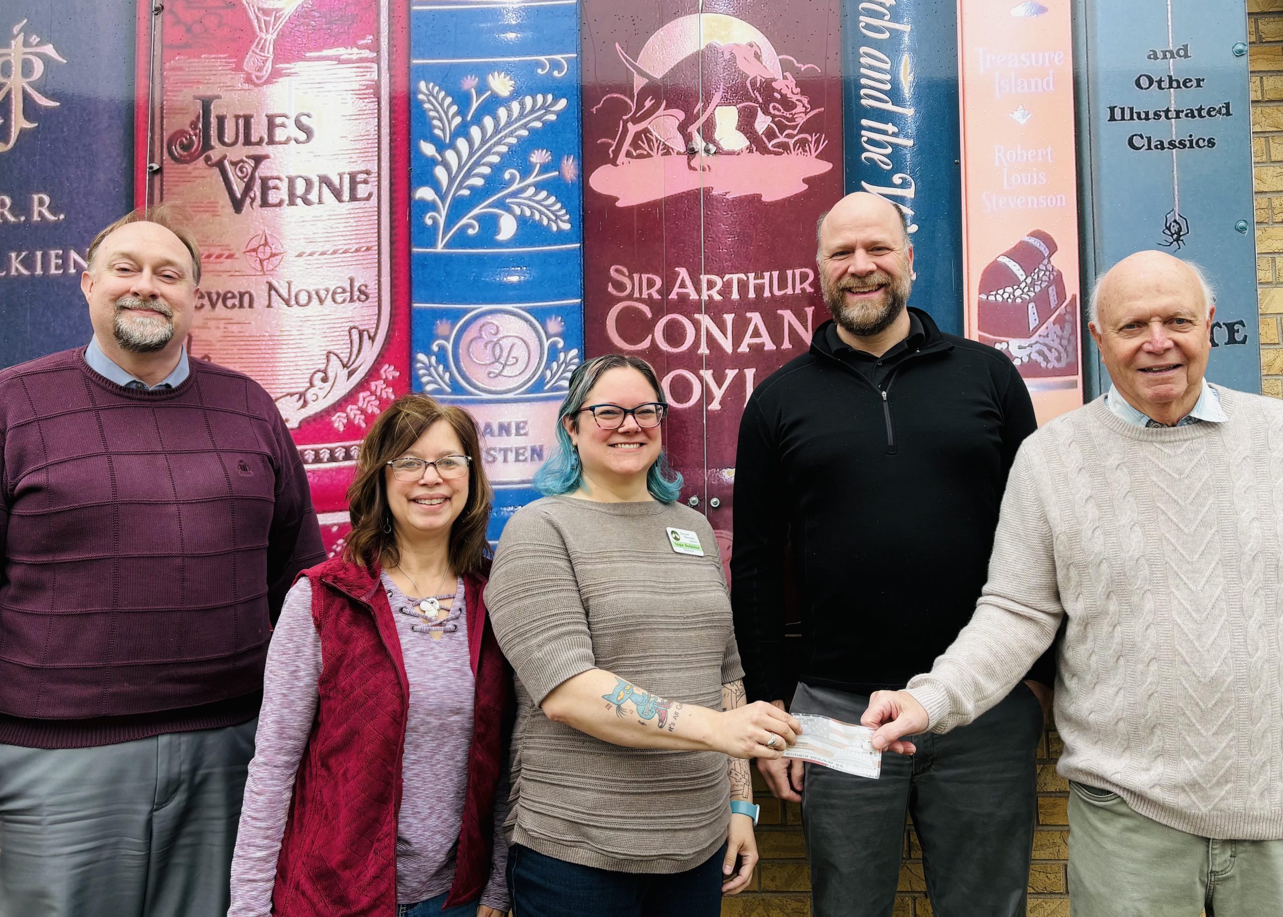 City Administrator Nick Koverman, Library Board President Audrey Kevan, Library Director Angie Bodzislaw, and the Schmitz family—Ted and Dave—are featured in this photo. The Schmitz family is presenting a check to the library to fund the purchase of the former fire hall property on Summit Street, ensuring the site's future use as a library.